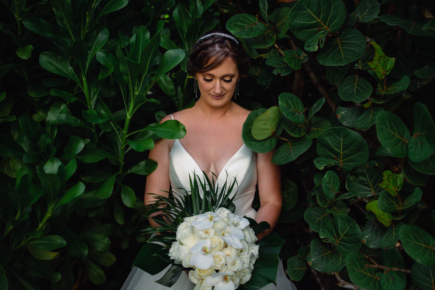 Bride in white gown holds elegant white bouquet, standing among lush green leaves in a serene outdoor setting.