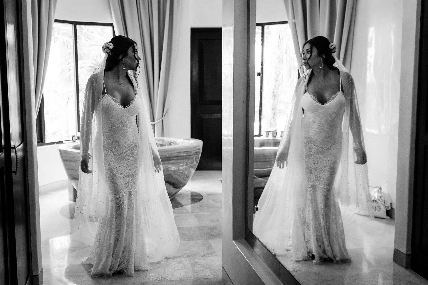 Bride in lace wedding gown and veil poses before mirror in elegant room, captured in black and white photography.