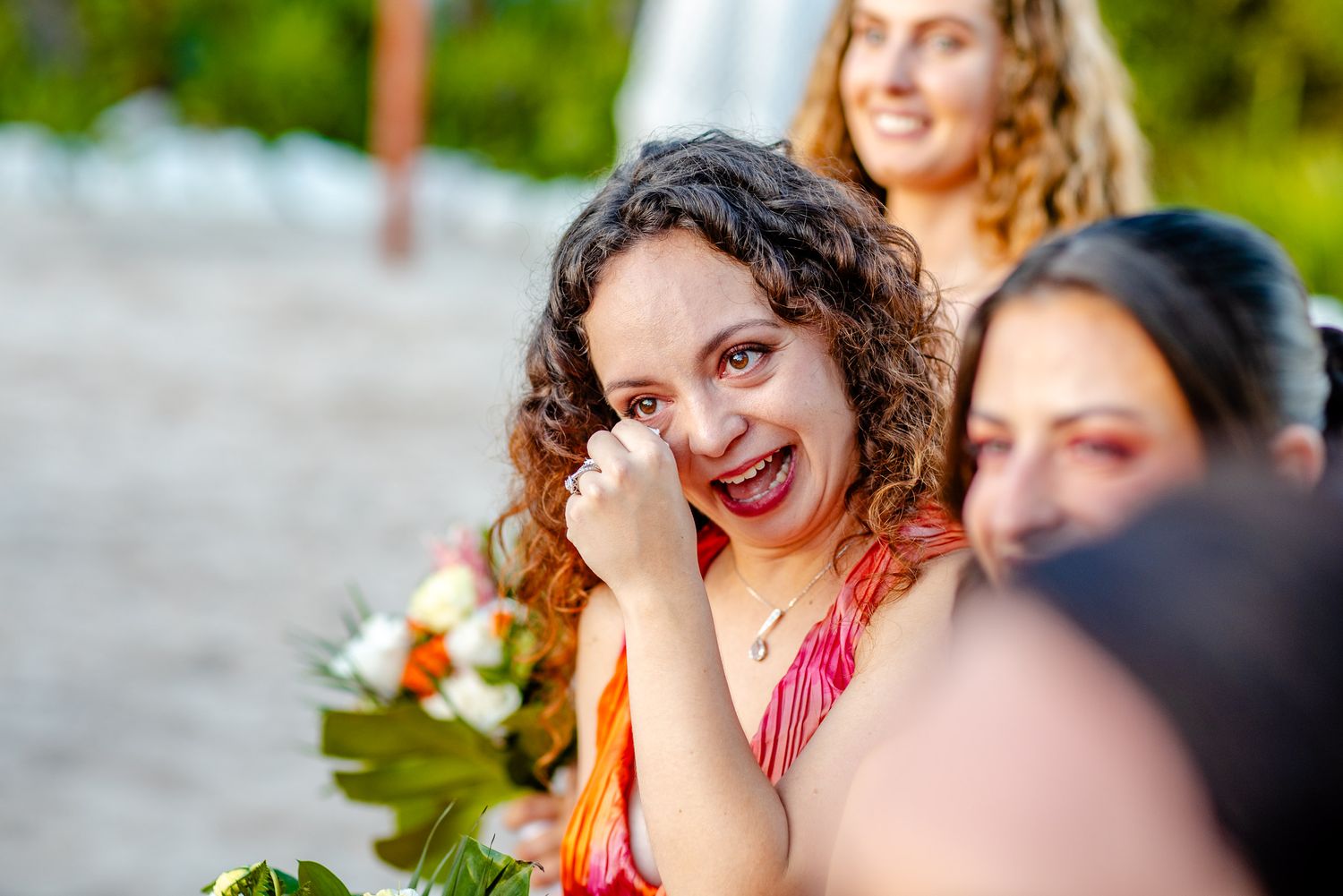 Emotional wedding guest wiping tears of joy at a beach ceremony, holding colorful bouquet.