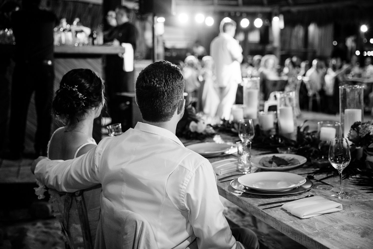 Black and white photo of bride and groom seated at wedding reception table, watching a speaker on stage.