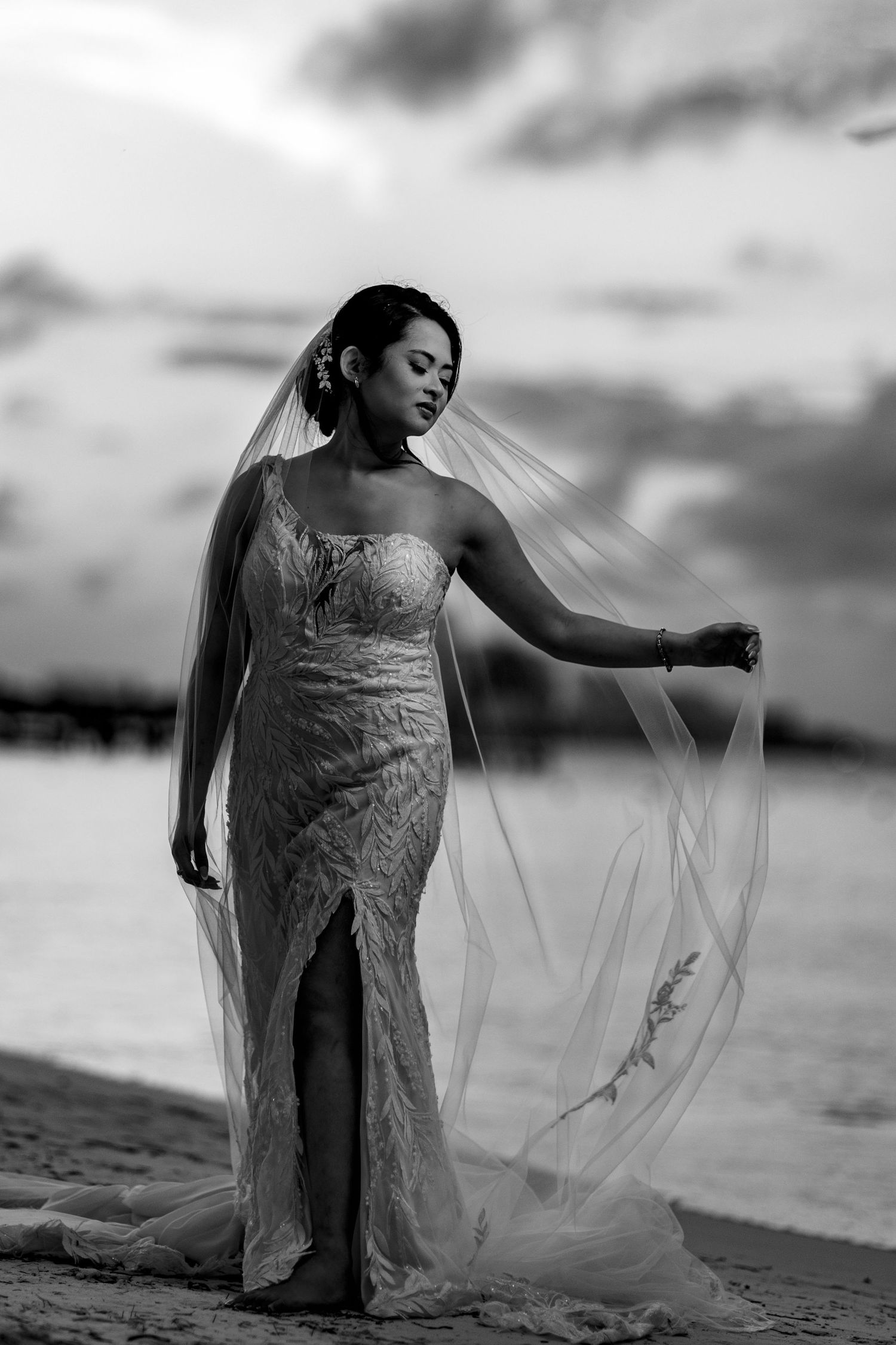 Black and white portrait of bride in lace gown holding veil on sandy beach at dusk.