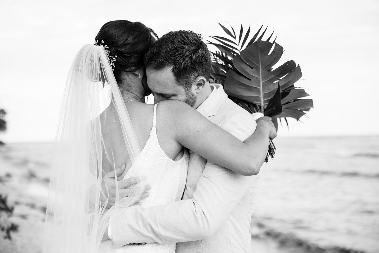 Black and white photo of bride and groom embracing on beach, bride holding tropical bouquet with palm leaves.
