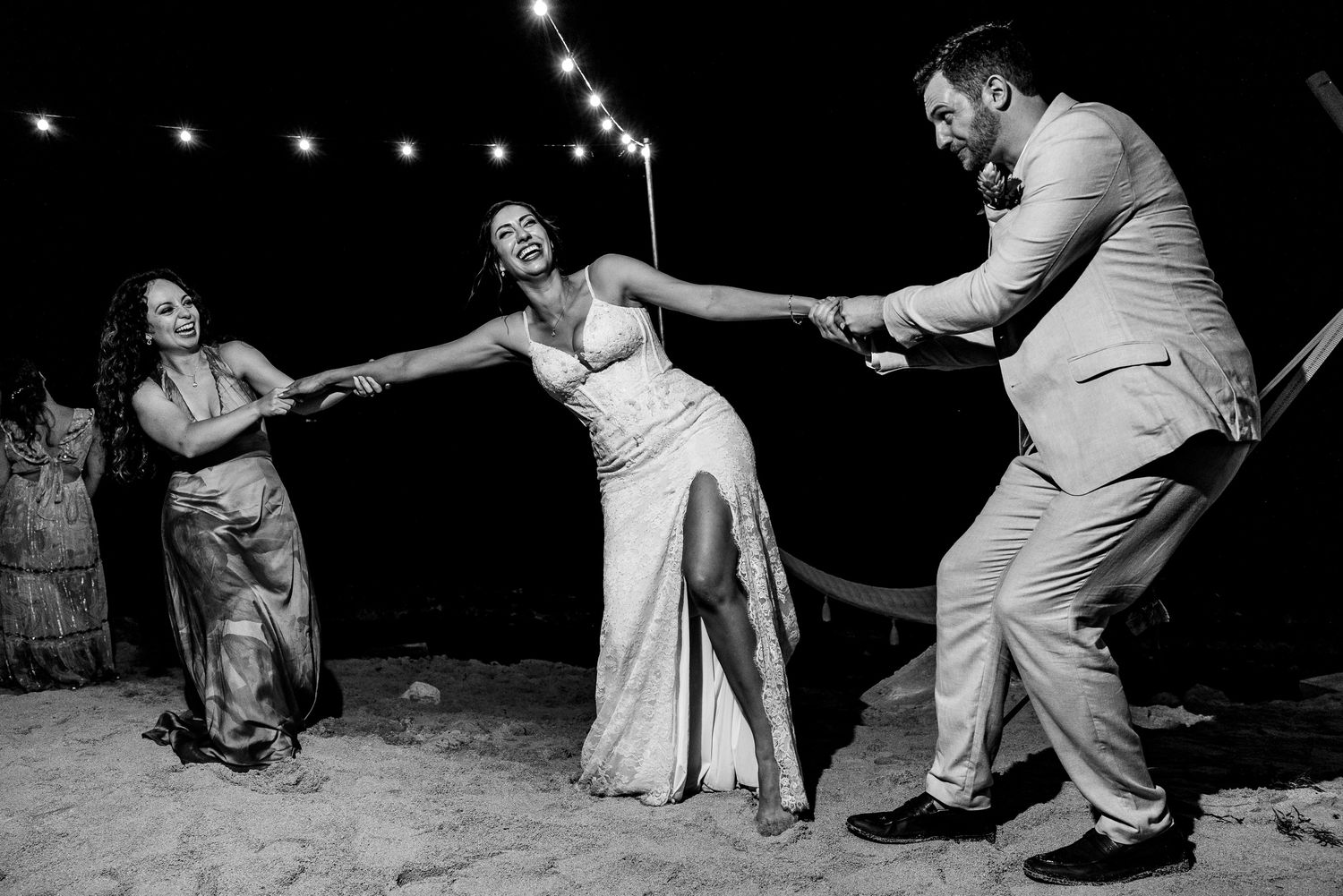 Bride and groom dance joyfully with a guest on a sandy beach at night, string lights glowing above.