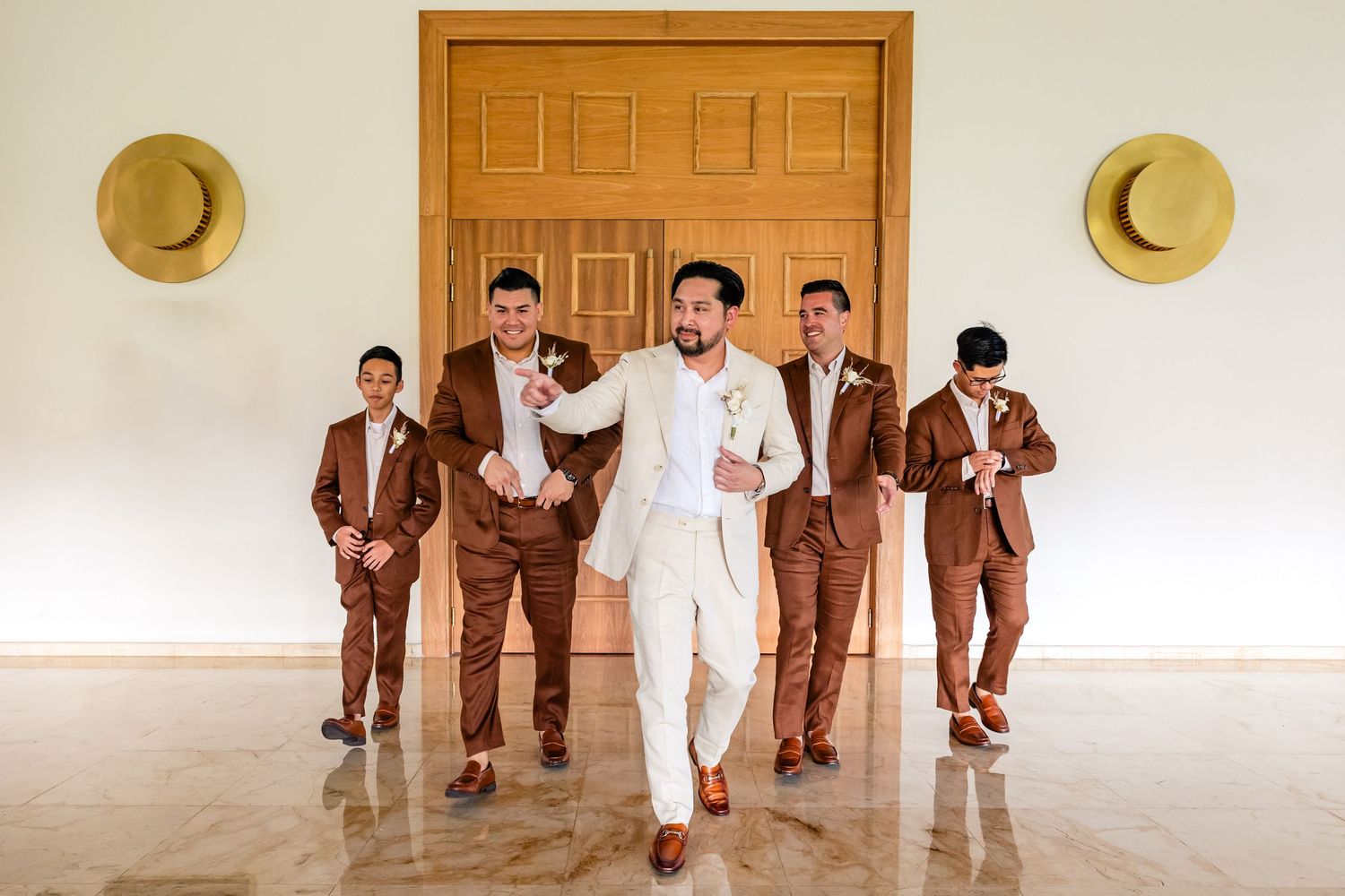 Groom in white suit walks confidently with groomsmen in brown suits inside an elegant venue with marble floors.