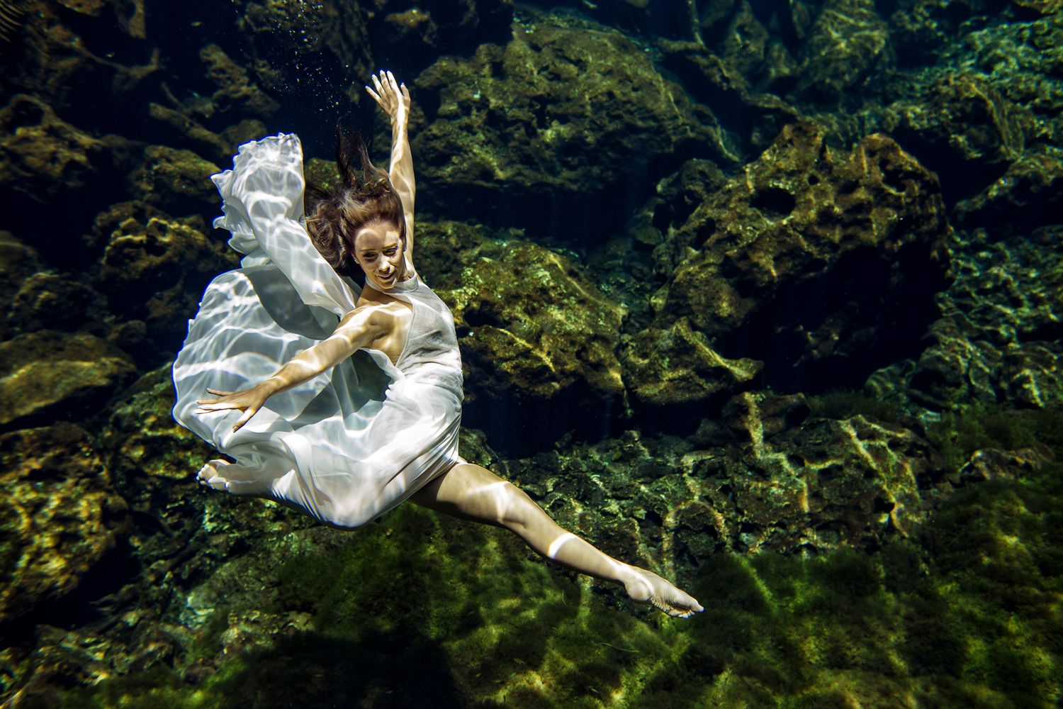 A woman in a flowing white dress gracefully poses underwater, surrounded by rocky green algae-covered walls.