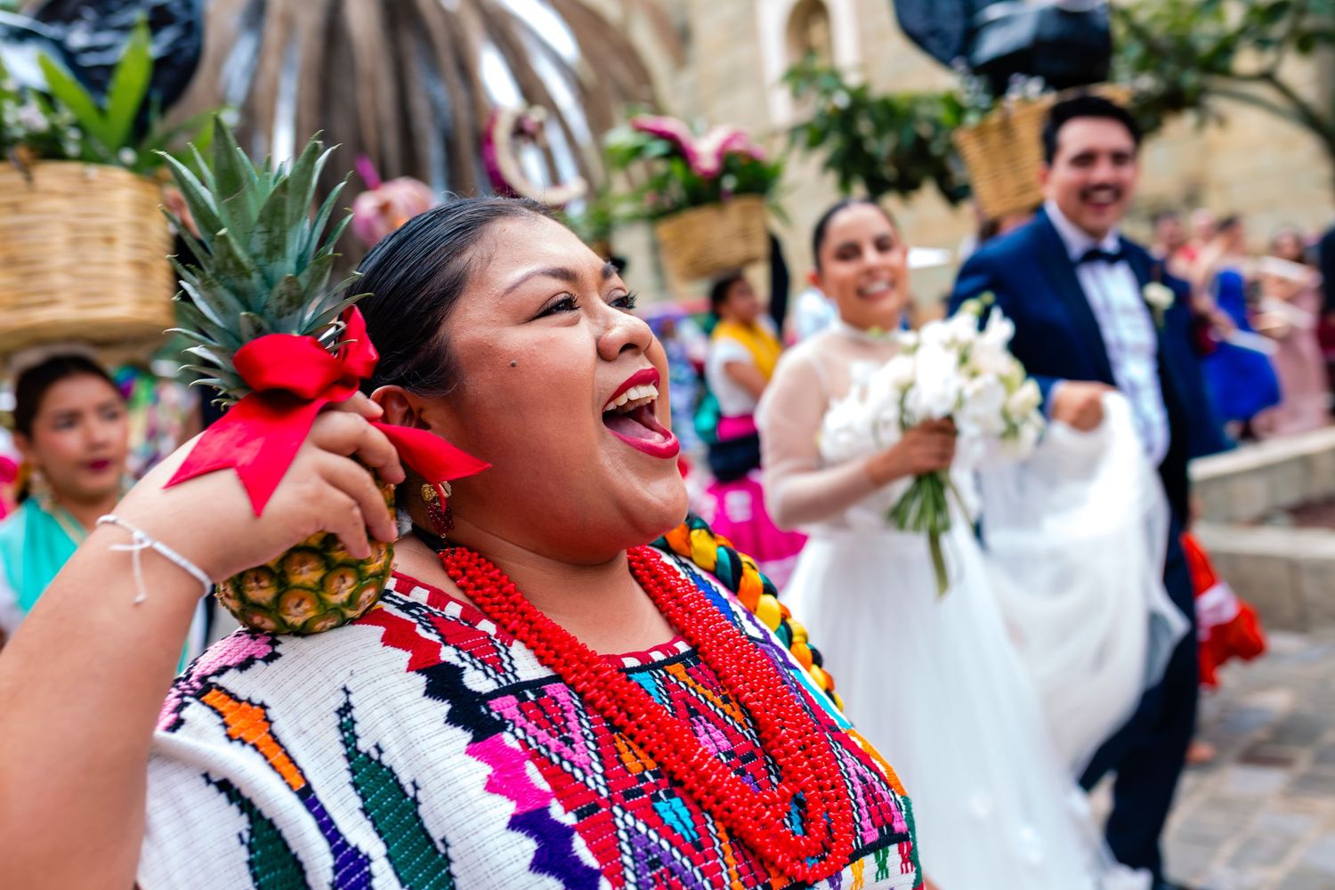 Joyful woman in colorful traditional Mexican dress celebrating at a wedding with bride and groom in background.