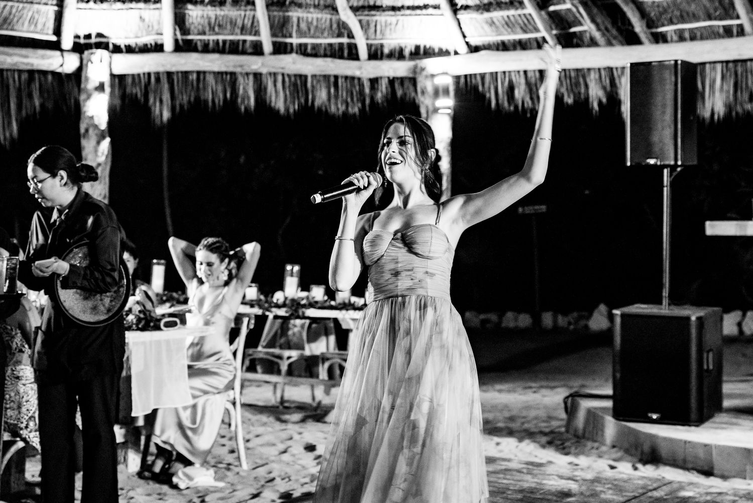 Woman singing into microphone at beach wedding reception under palapa in black and white.
