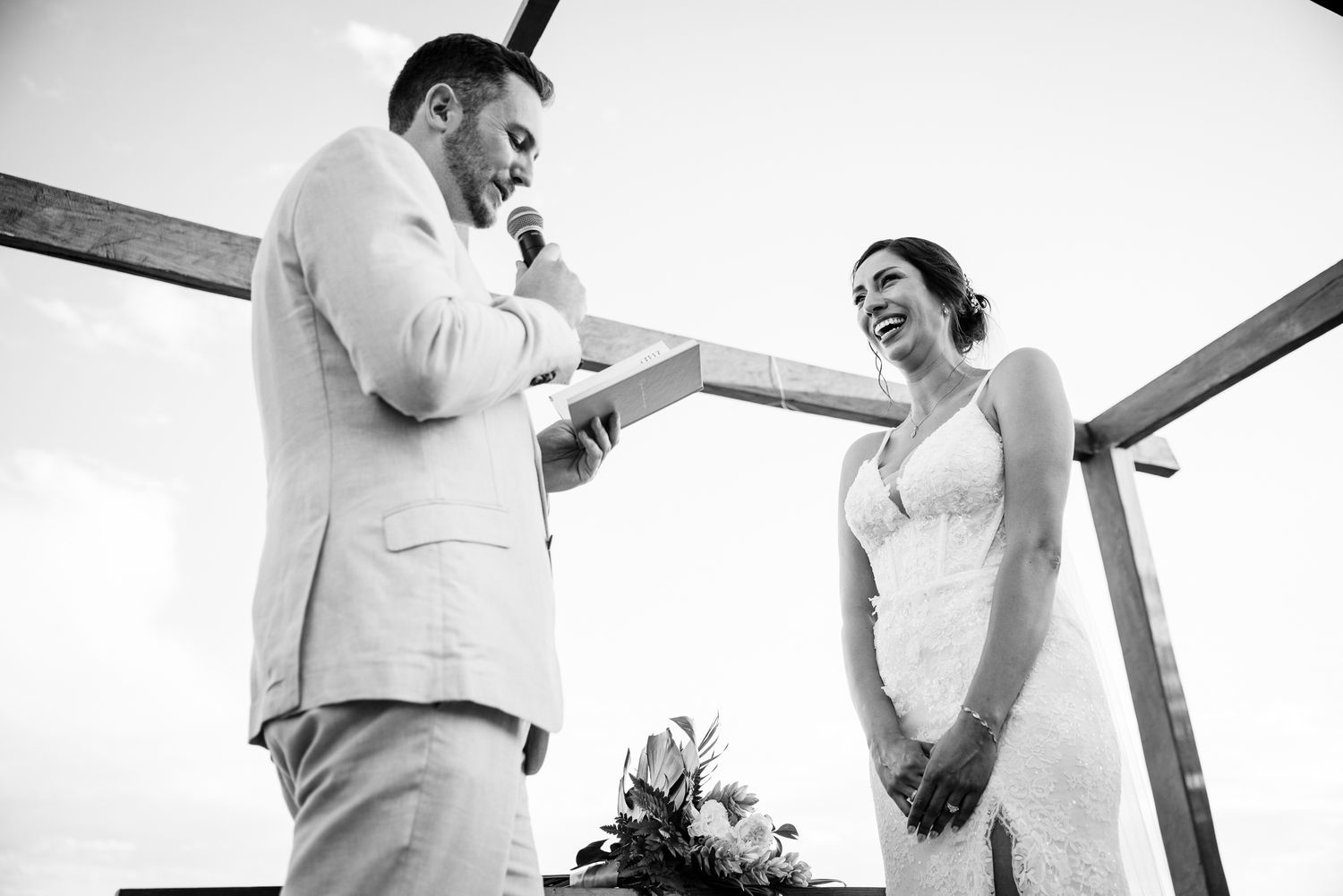 Black and white photo of groom reading vows to smiling bride at outdoor wedding ceremony with cross backdrop.