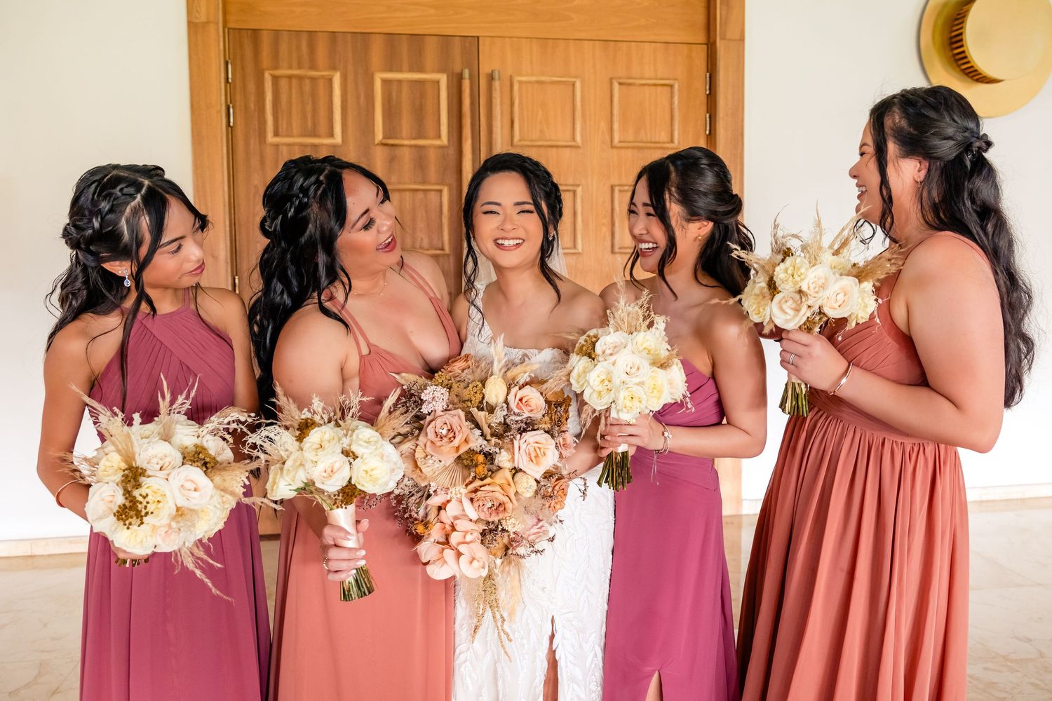 Bride and bridesmaids in pink dresses laughing together holding white and blush floral bouquets at a wedding.