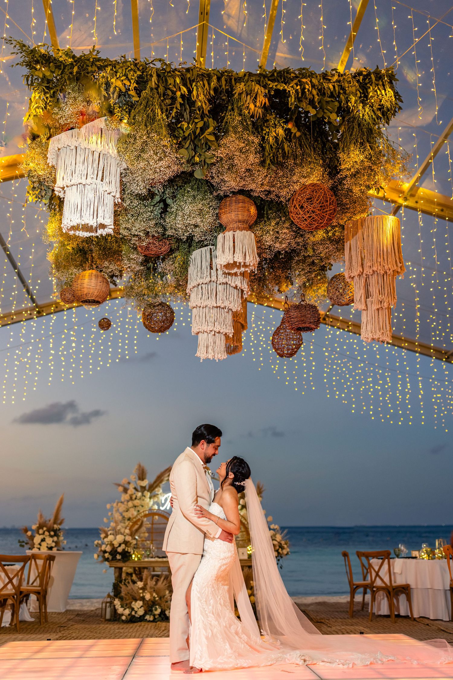 Couple shares first dance under stunning floral canopy with macrame chandeliers and fairy lights at beach wedding.