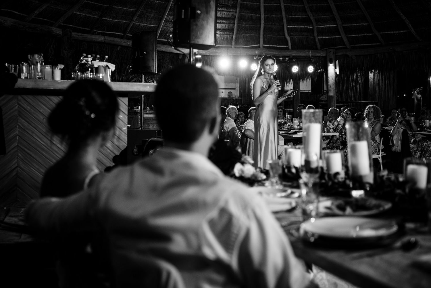 Black and white wedding reception photo of a woman giving a toast speech while guests listen at candlelit tables.