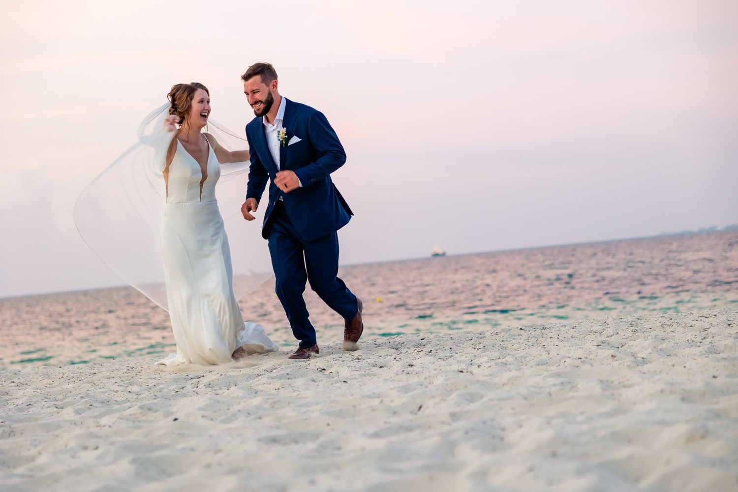 Bride and groom walking hand in hand on a sandy beach at sunset in wedding attire.