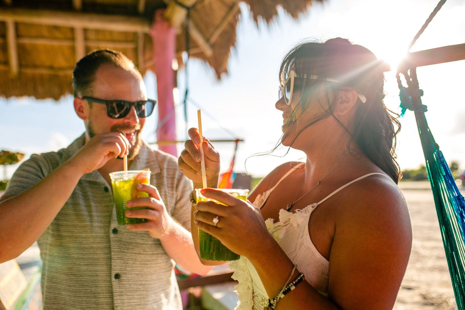 Couple enjoying tropical green drinks with straws at a sunny beach bar, smiling and relaxed on vacation.