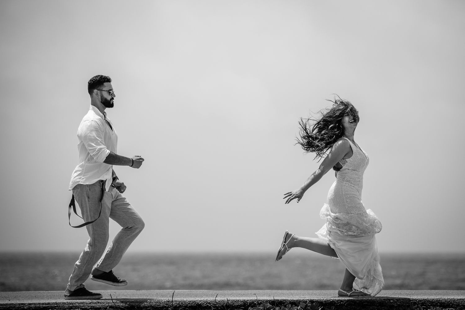 Black and white photo of a couple running joyfully by the ocean, woman in white dress, man in white shirt.