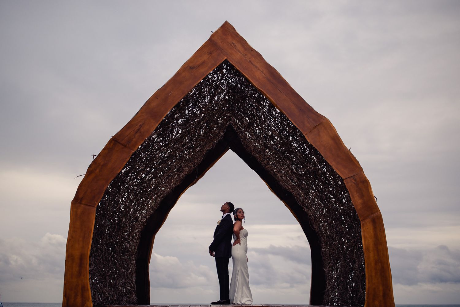 Bride and groom stand kissing under ornate arch sculpture on cloudy day at outdoor wedding ceremony.