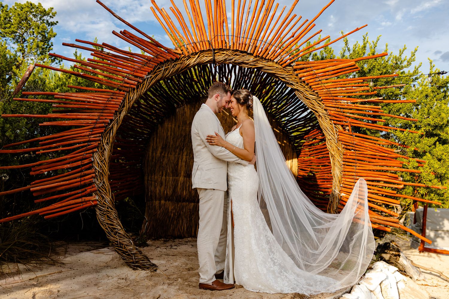 Bride and groom kissing under a stunning sunburst wicker arch sculpture at an outdoor wedding venue.