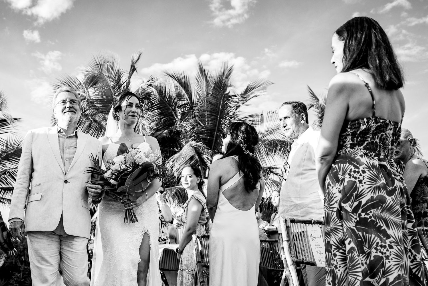 Black and white outdoor wedding ceremony with couple holding bouquet, guests, and palm trees in tropical setting.