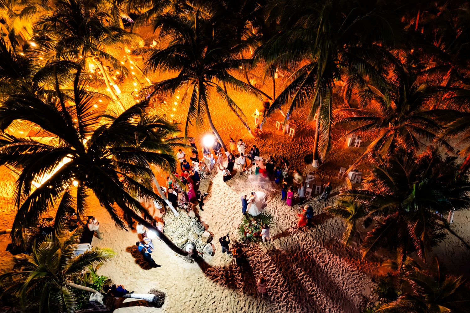 Aerial night view of a tropical beach party under palm trees with warm orange lighting and a crowd of guests.