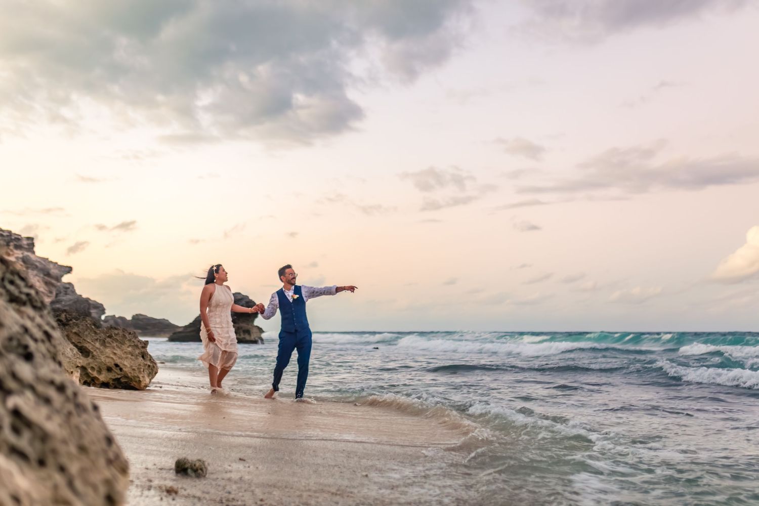 Couple in wedding attire walking on a rocky beach at sunset, groom pointing toward the ocean waves.