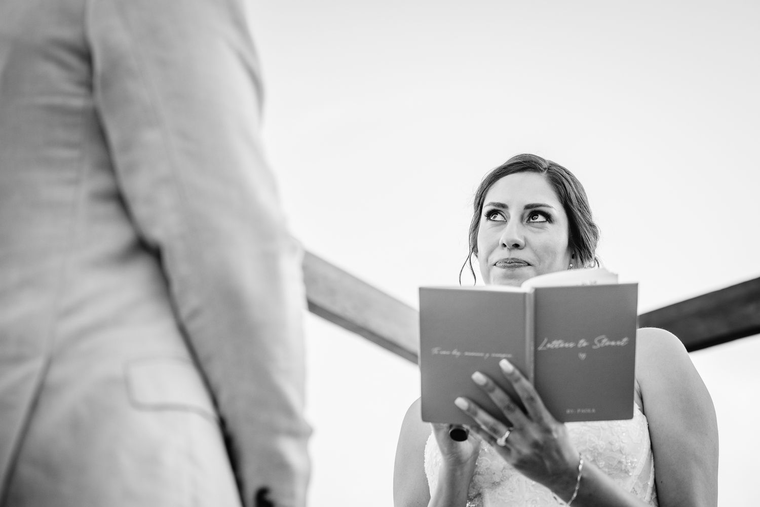 Bride reads vows from booklet while looking up at groom during black and white wedding ceremony photo.