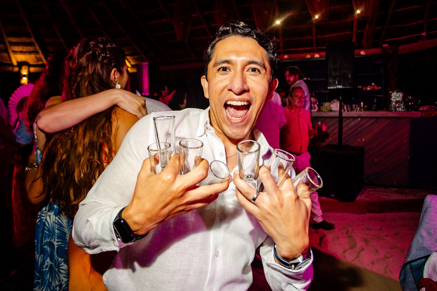 Excited man holding multiple shot glasses at a lively party celebration.