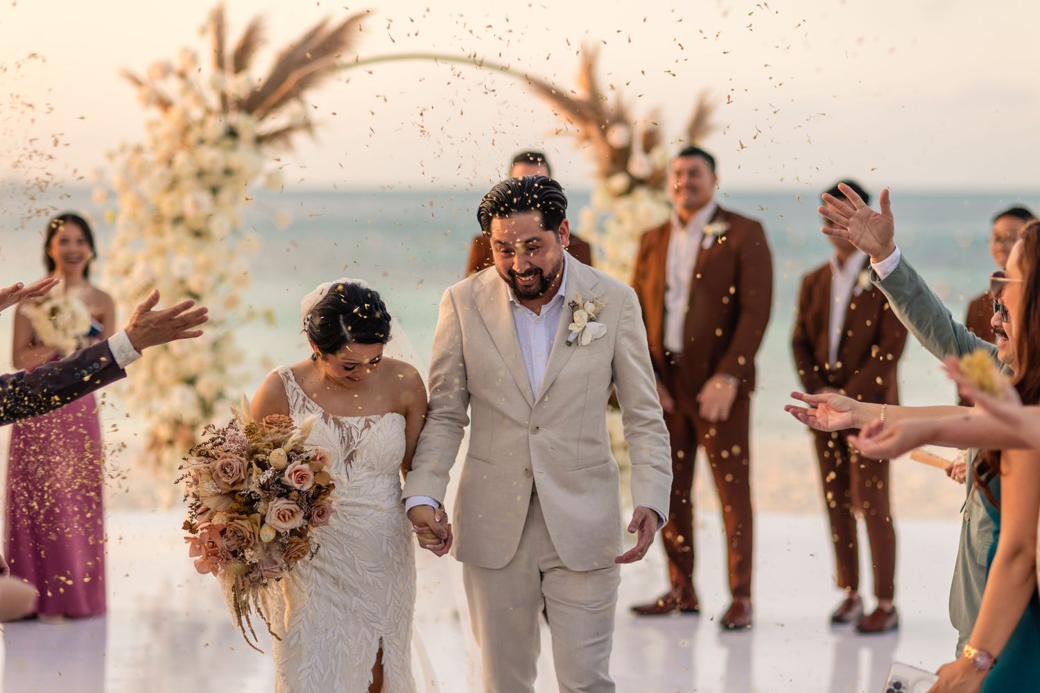 Joyful newlyweds walk through confetti shower at beachside wedding ceremony with guests celebrating around them.