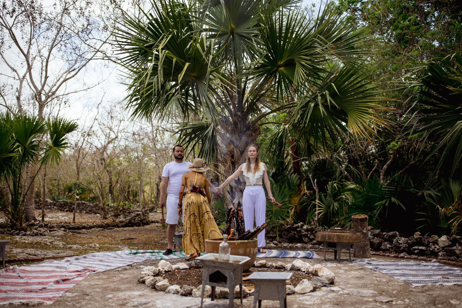 Two people stand in a tropical outdoor ceremony space with palm trees, a fire pit, and a traditionally dressed officiant.