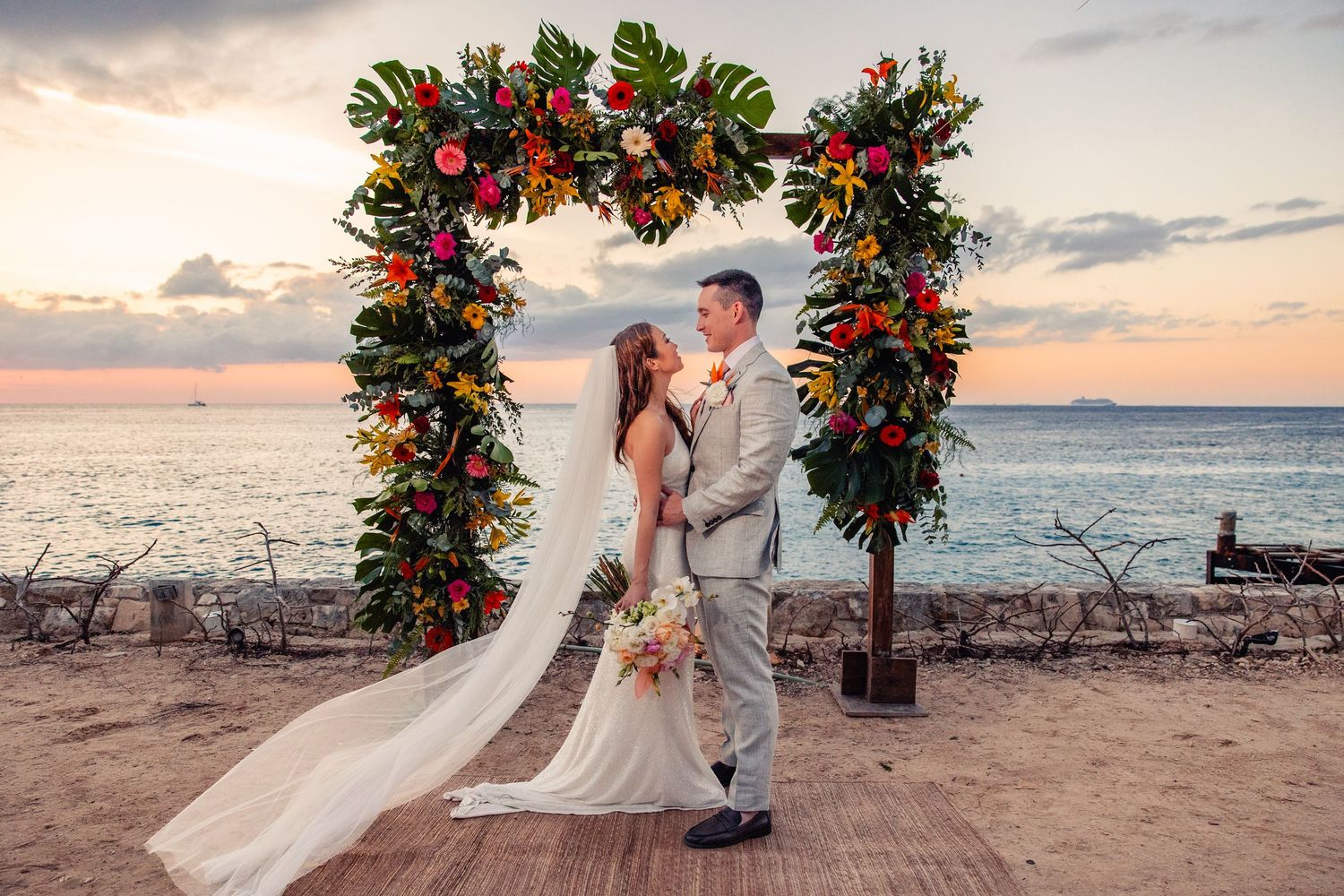 Tropical beach wedding ceremony setup with red and green floral arrangements during a beautiful sunset over the ocean.