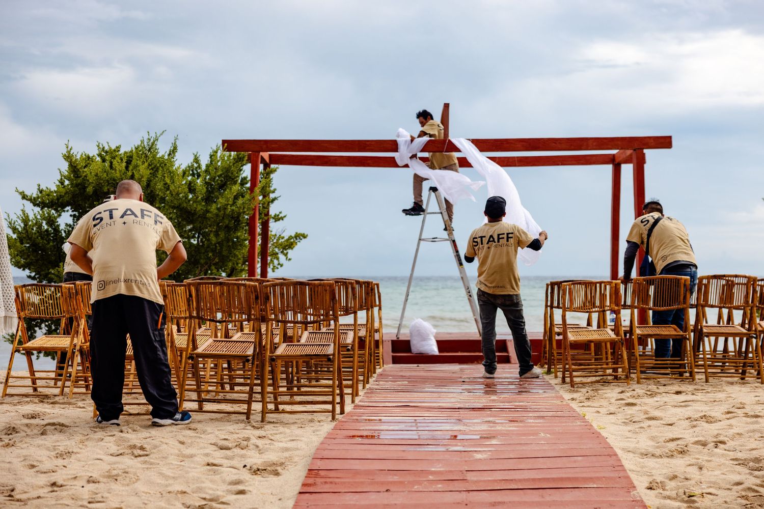 Staff members setting up a beachside wedding ceremony with red arch, white draping, and wooden chairs on sandy shore.