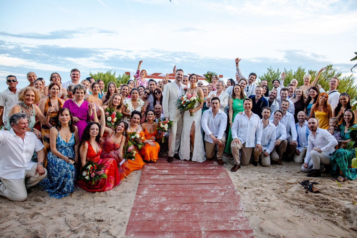 Large wedding party group photo on sandy beach at sunset with bride and groom at center.
