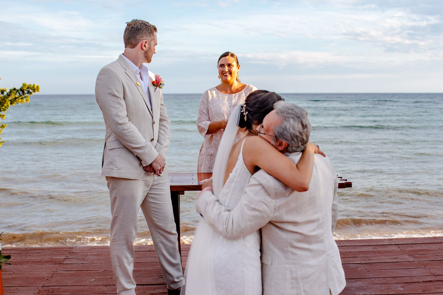 Bride and groom share an emotional hug with a guest during a beachside wedding ceremony.