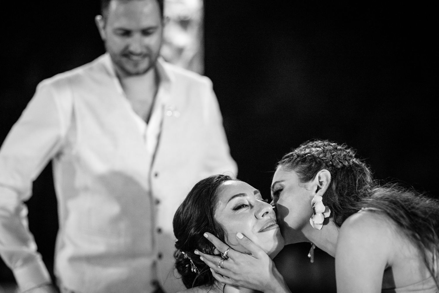 Black and white photo of a bride receiving a kiss on the cheek from a bridesmaid at a wedding celebration.
