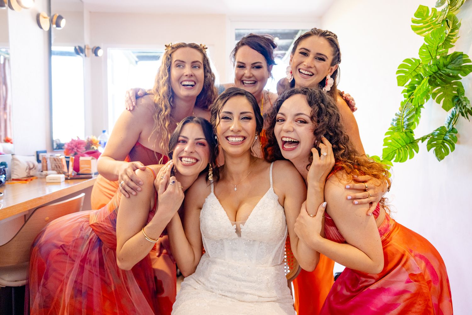 Bride in white gown poses joyfully with five bridesmaids in red and orange dresses in a bright bridal suite.