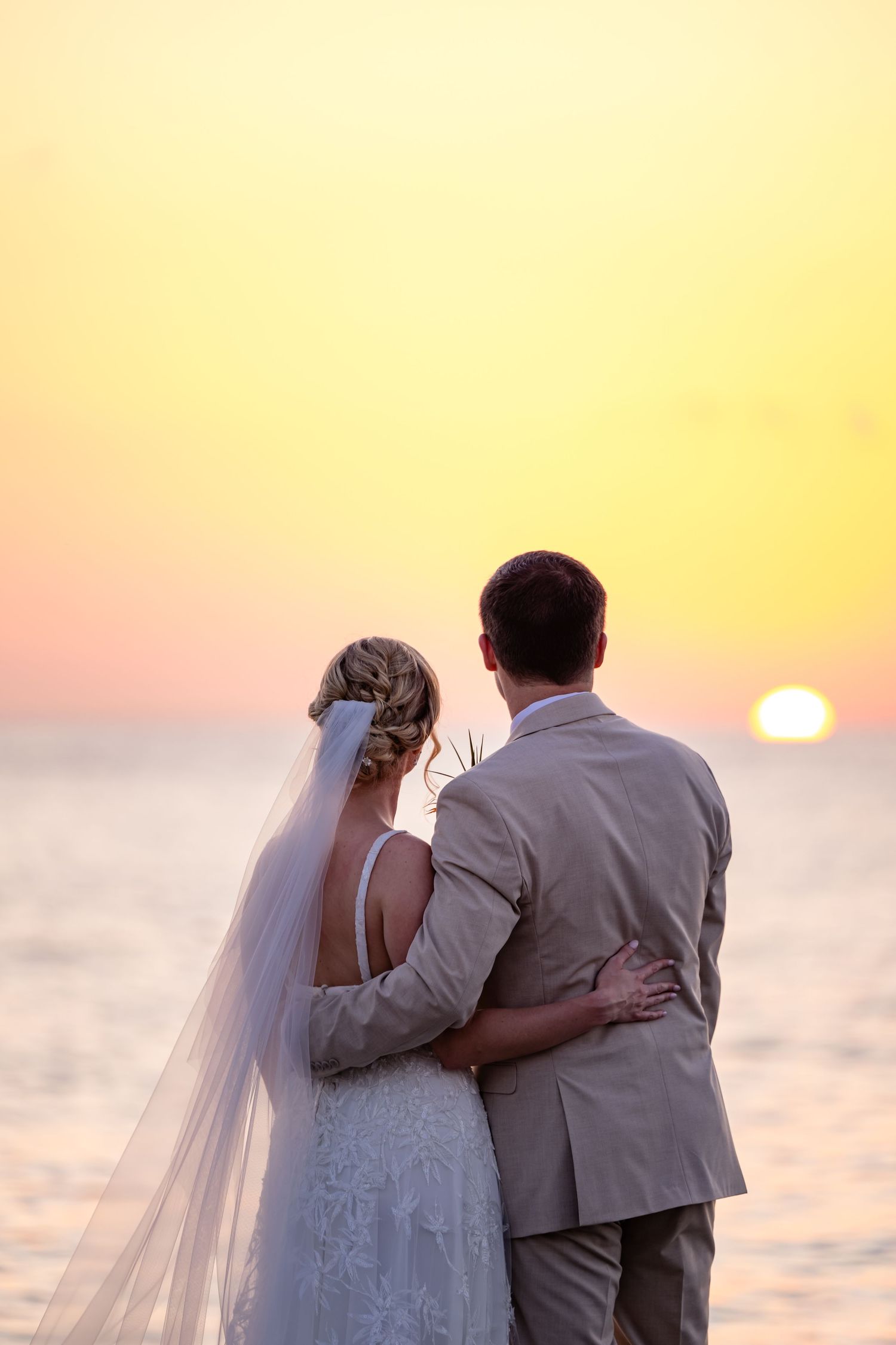 A couple embraces while watching a beautiful sunset over the ocean on their wedding day.