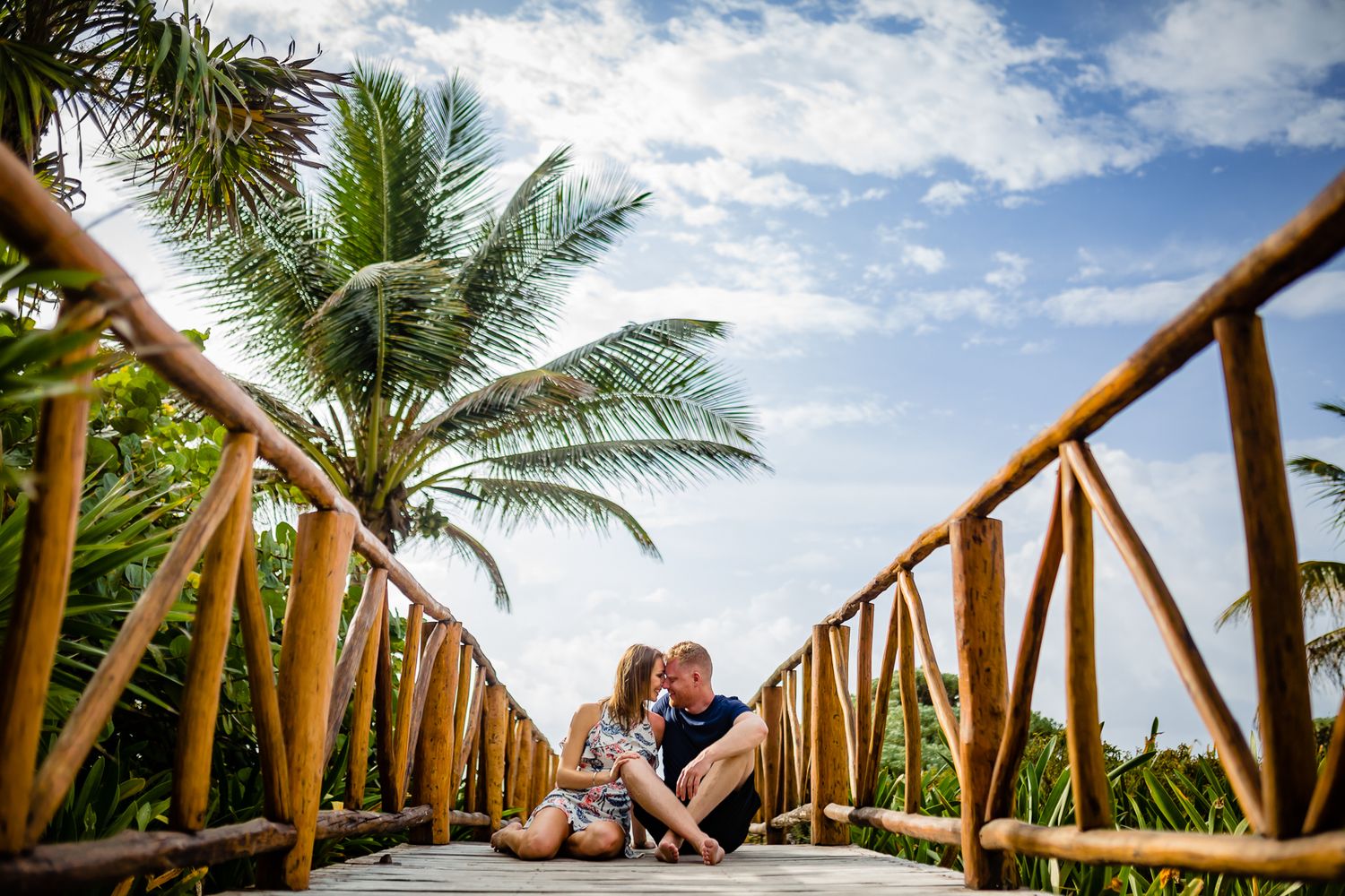 Couple shares romantic moment under wooden walkway surrounded by tropical palm trees and blue sky.