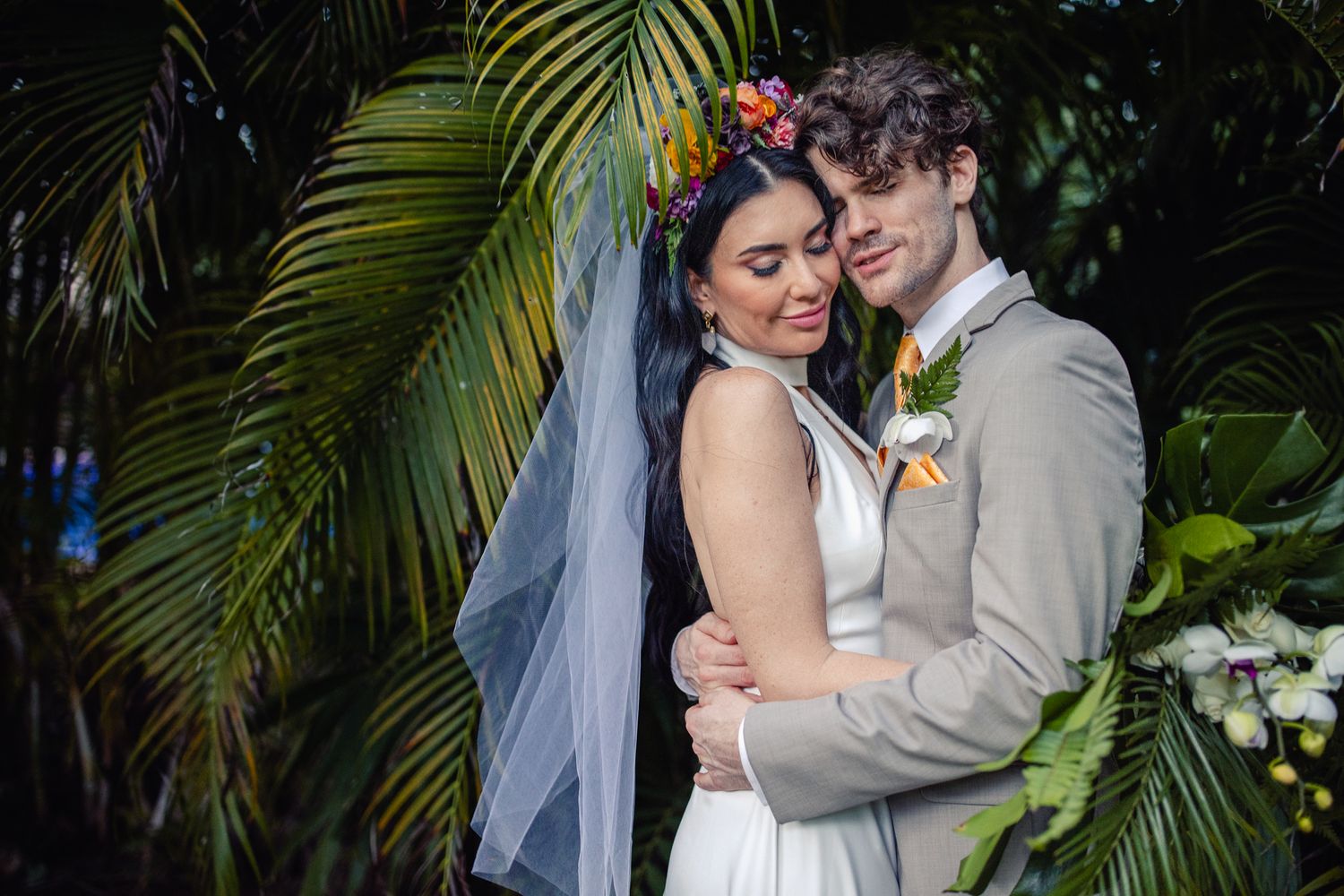 Bride and groom embrace tenderly among tropical palm leaves, bride wearing floral crown and white veil.