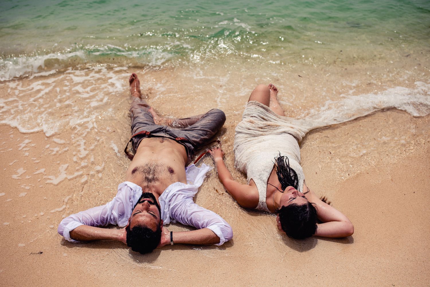 Two men lying on sandy beach shore with ocean waves, relaxing outdoors in casual summer attire.
