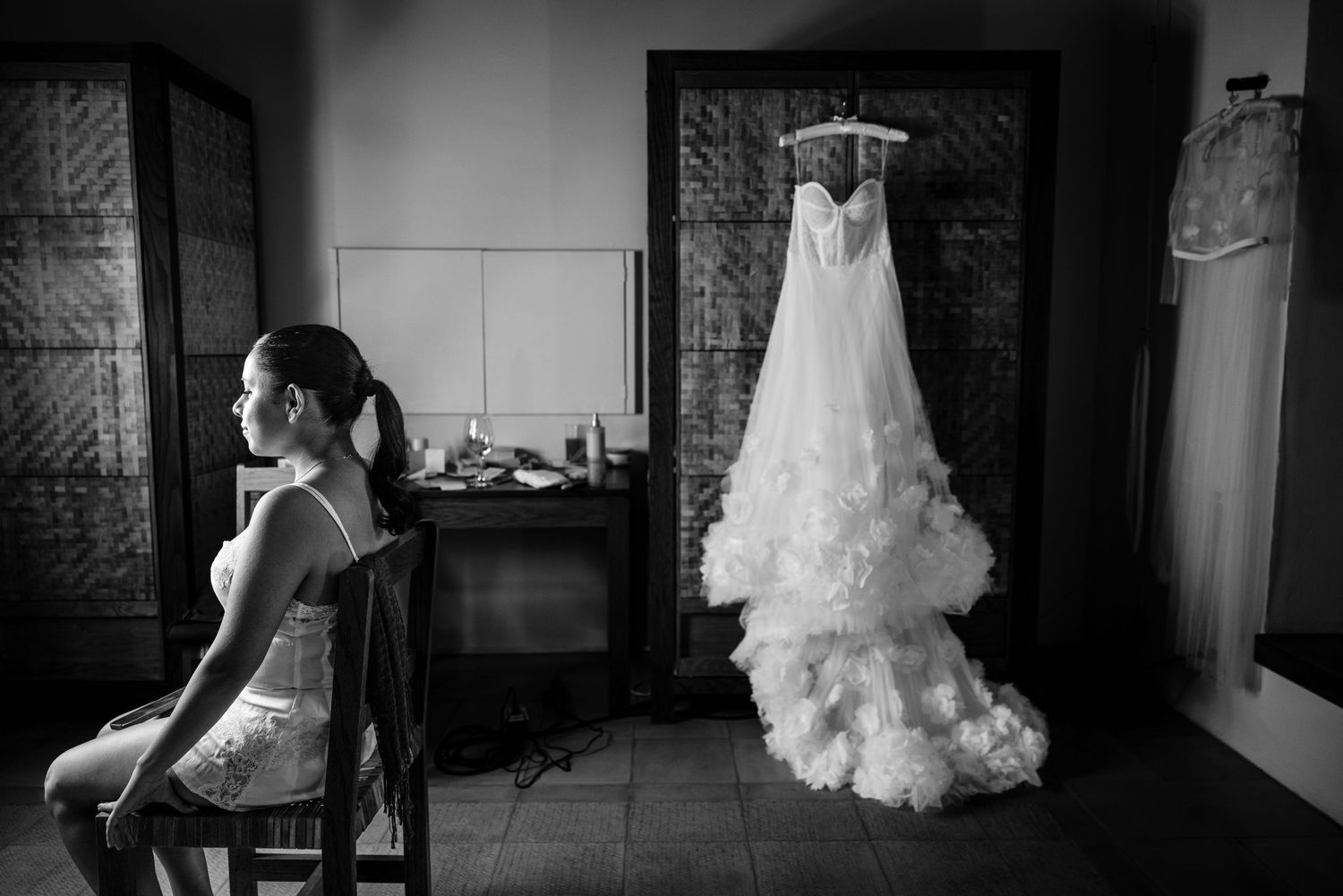Black and white photo of bride sitting in chair beside her elegant ruffled wedding gown hanging in a sunlit room.