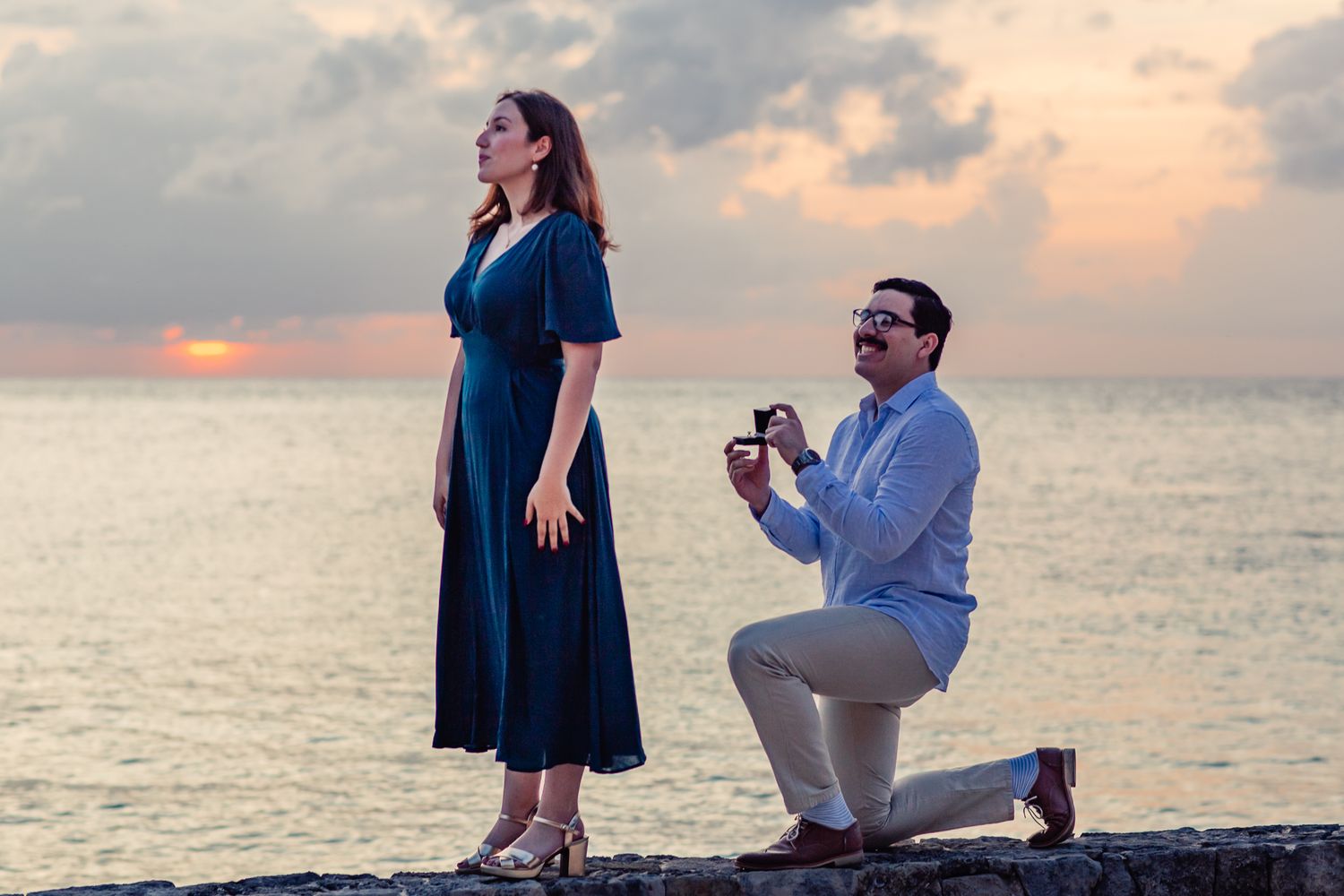 A romantic beach proposal at sunset with a couple silhouetted against the orange sky.