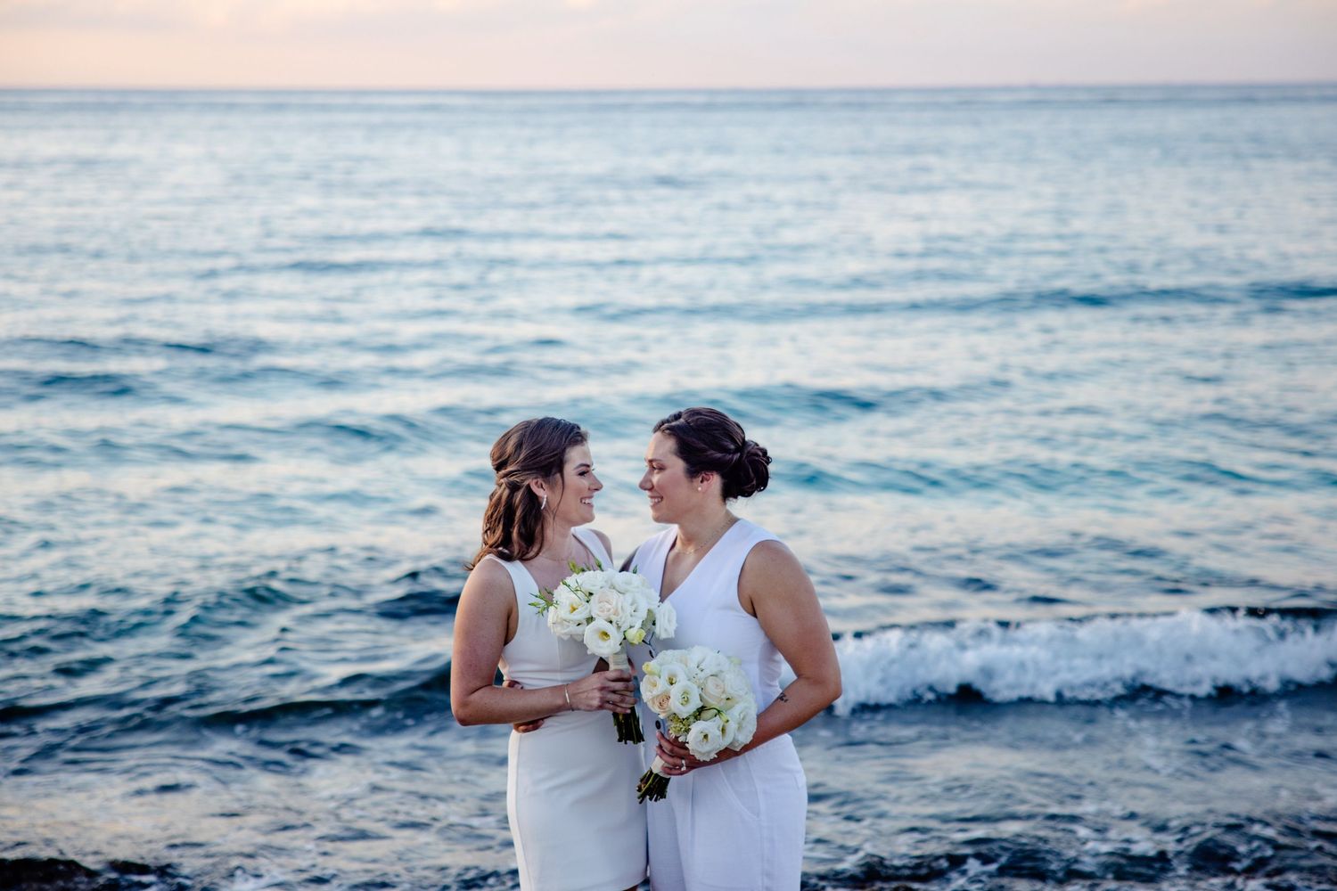 Two people in white dresses holding bouquets embrace on beach at sunset with ocean waves in background.
