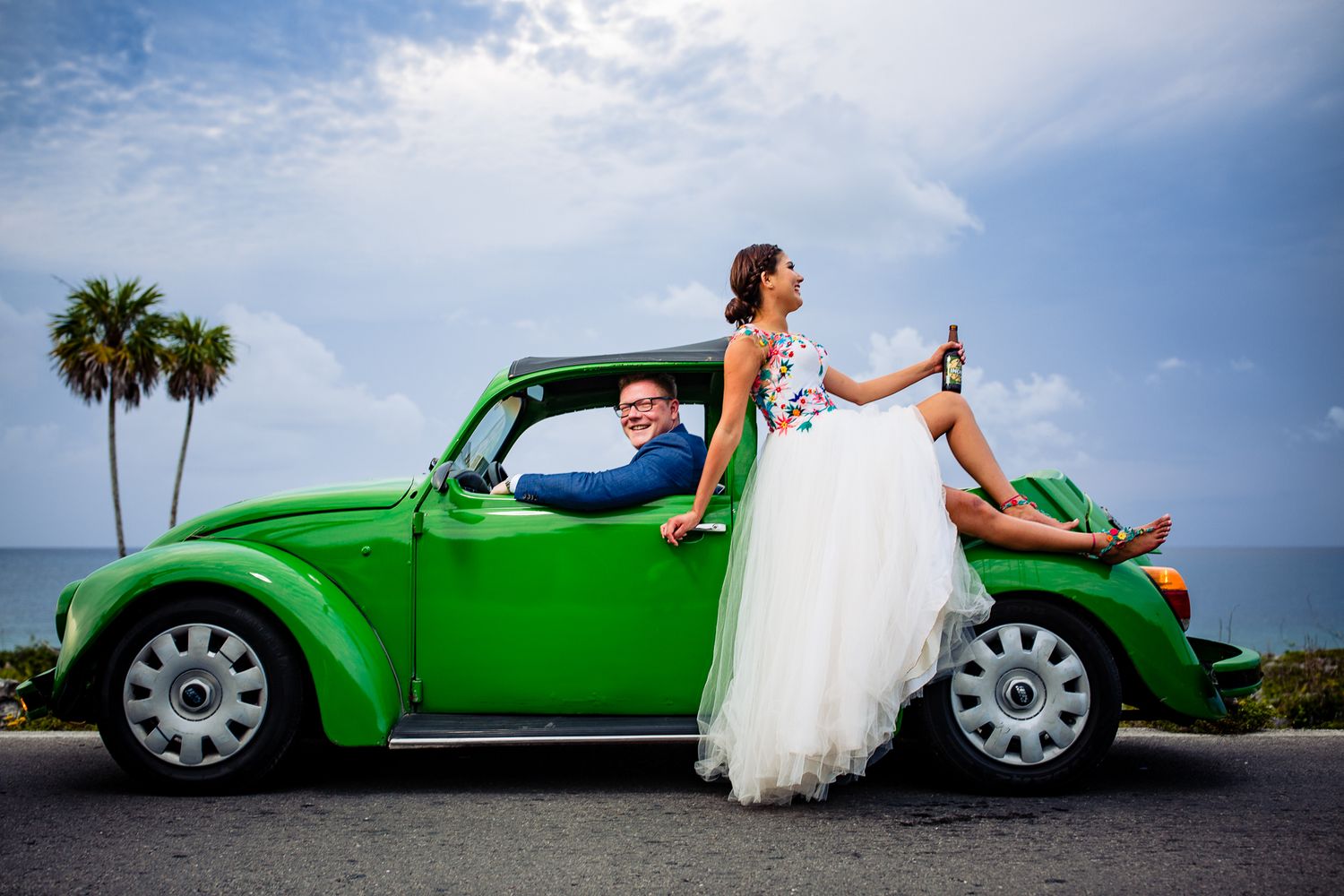 Bride sits on green VW Beetle hood holding champagne while groom drives, with palm trees and ocean backdrop.