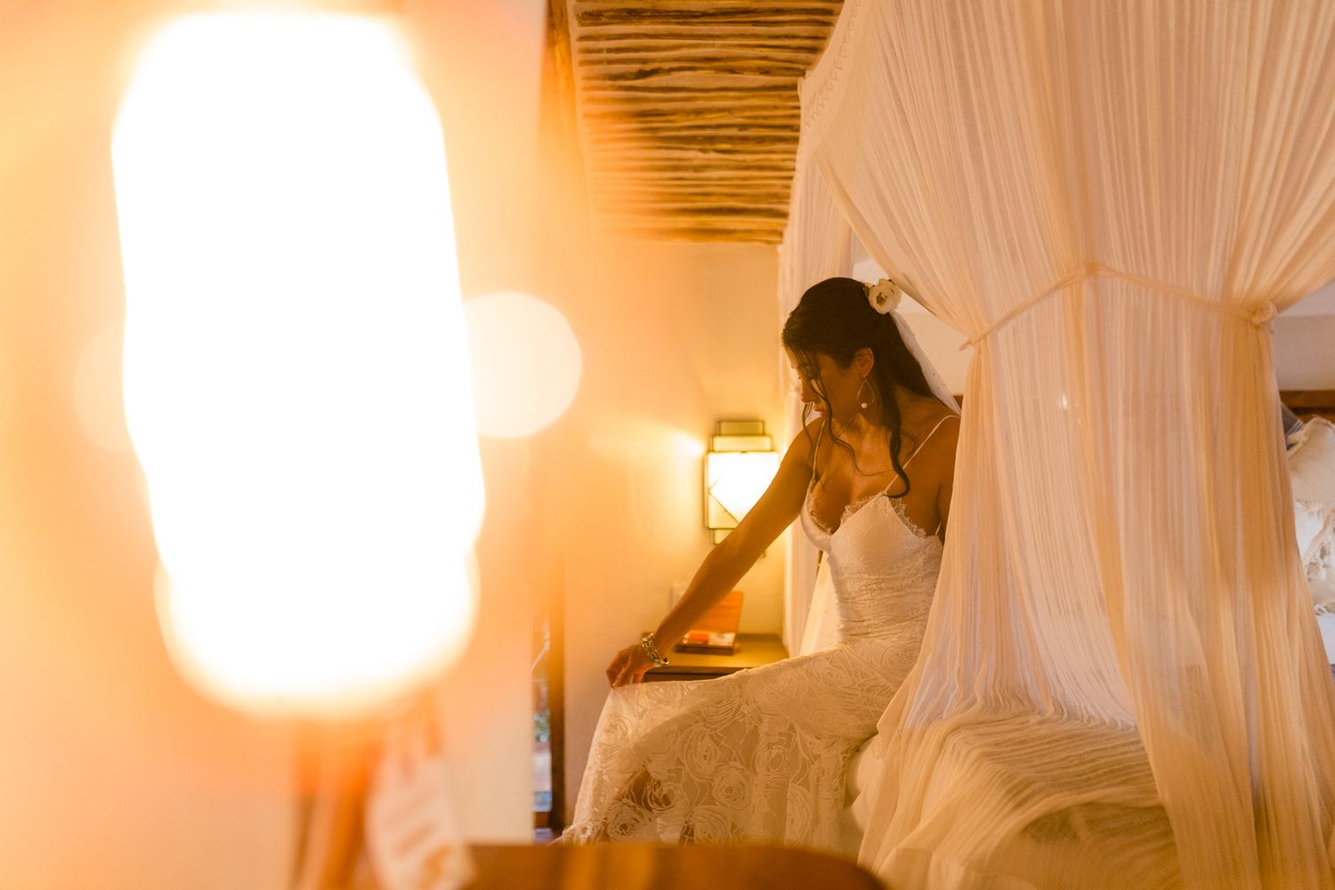 Bride in white wedding dress sitting on canopy bed in warmly lit rustic room.