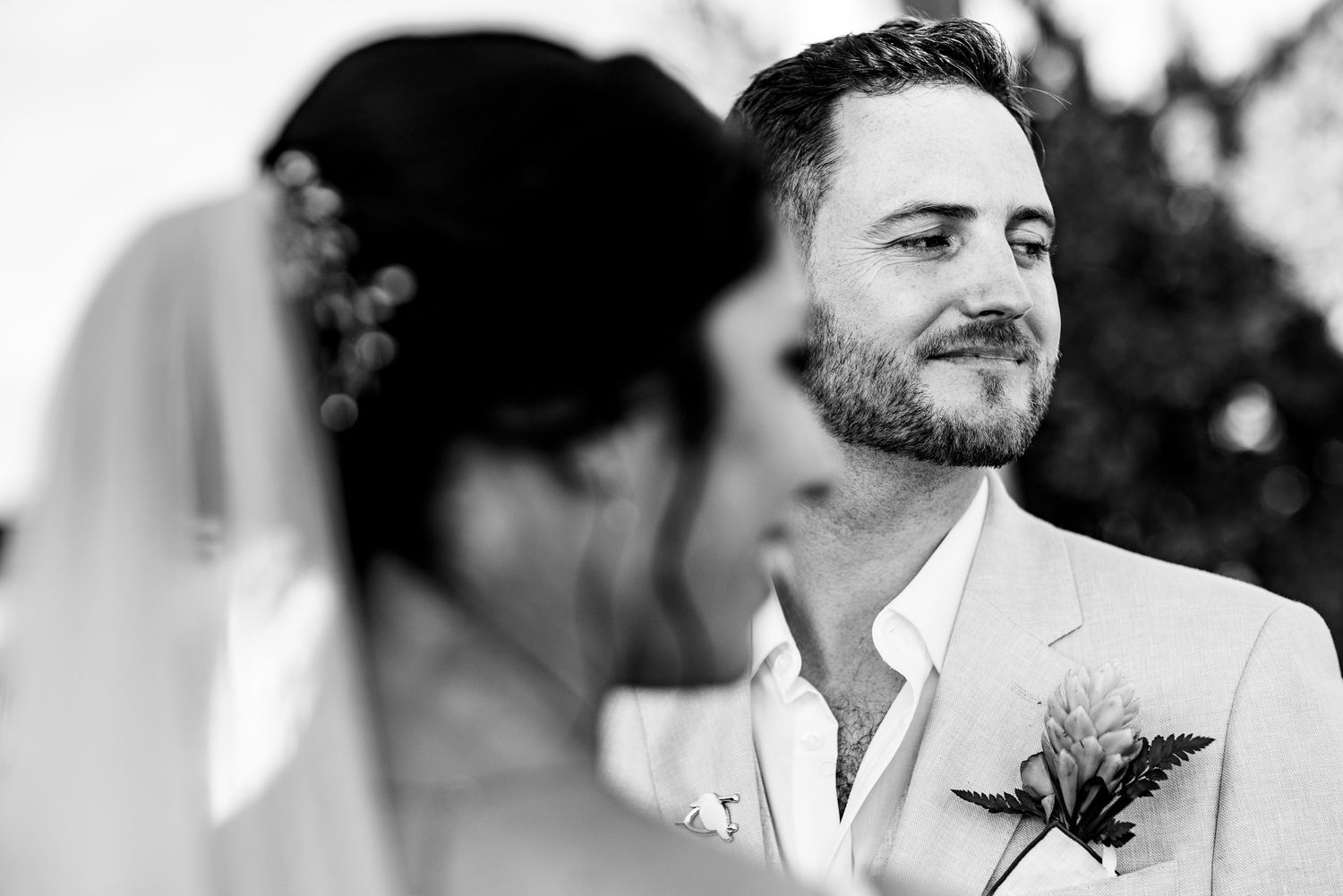 Black and white wedding photo of groom smiling at bride in veil, wearing suit with floral boutonniere.