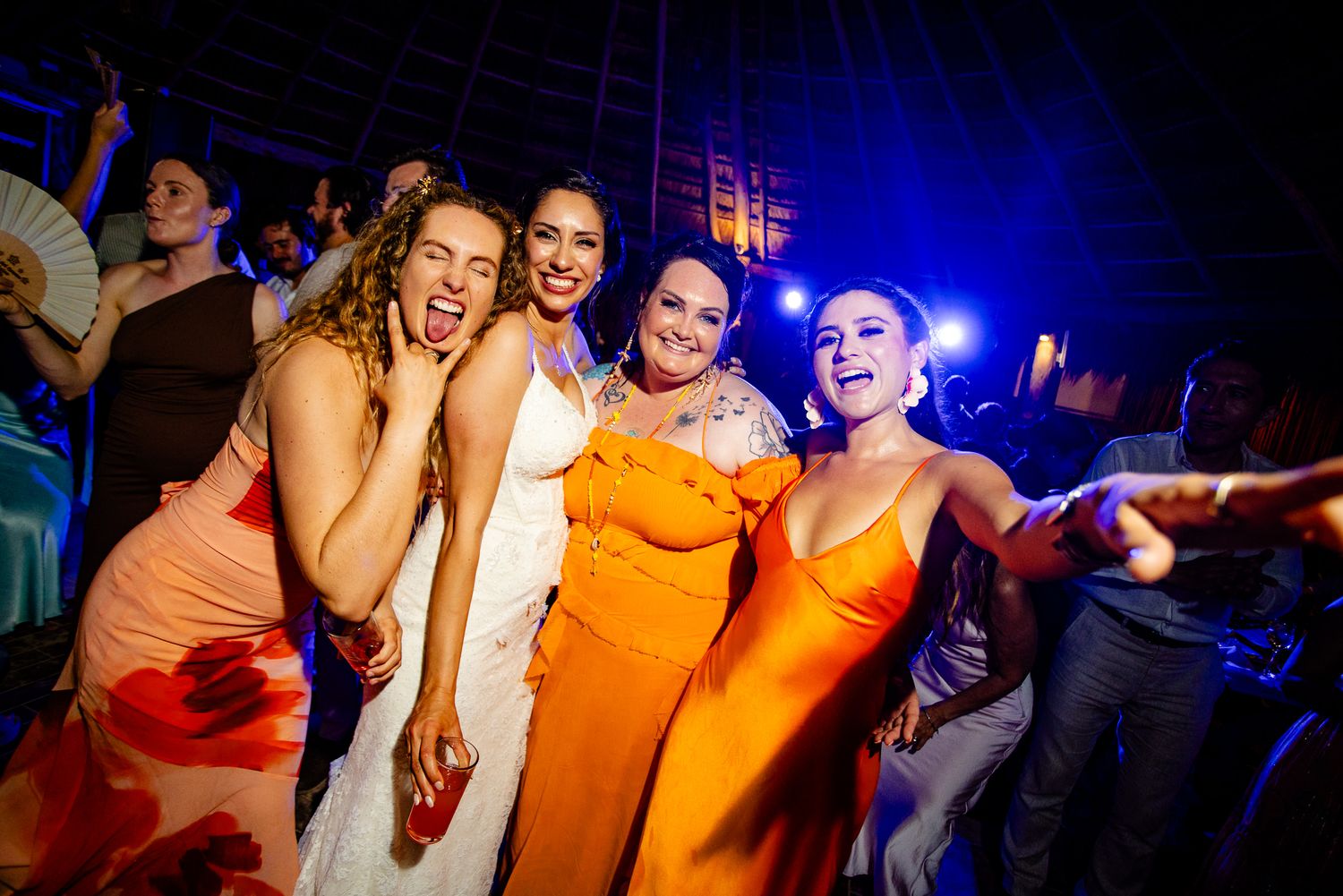 Four women in elegant dresses smile and dance together at a vibrant evening wedding reception.