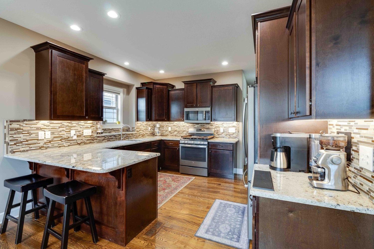 Modern kitchen design with dark cabinets, stone backsplash, and breakfast bar featuring hardwood floors and recessed lighting.