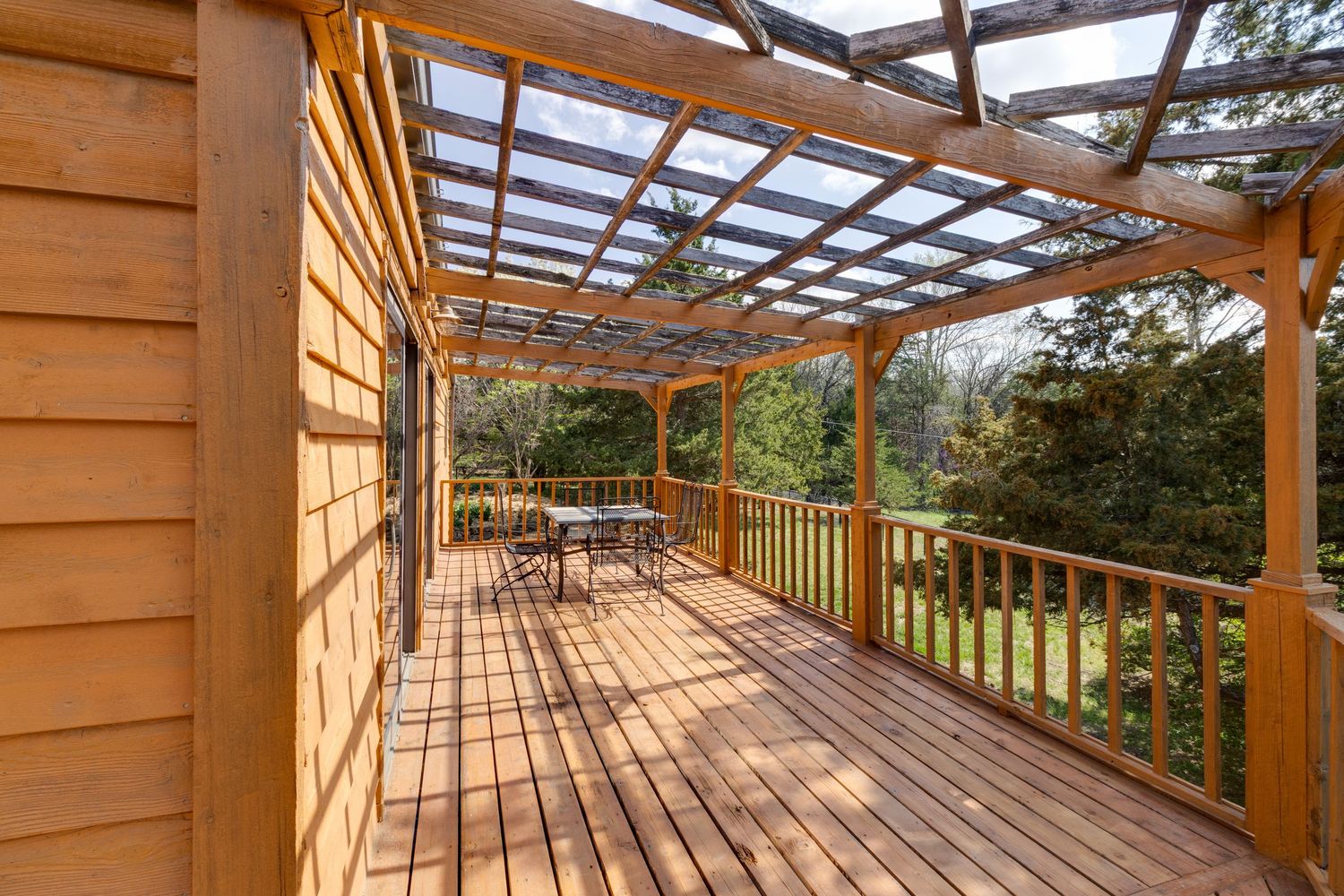 Wooden deck with pergola and railing overlooking natural scenery in warm sunlight.