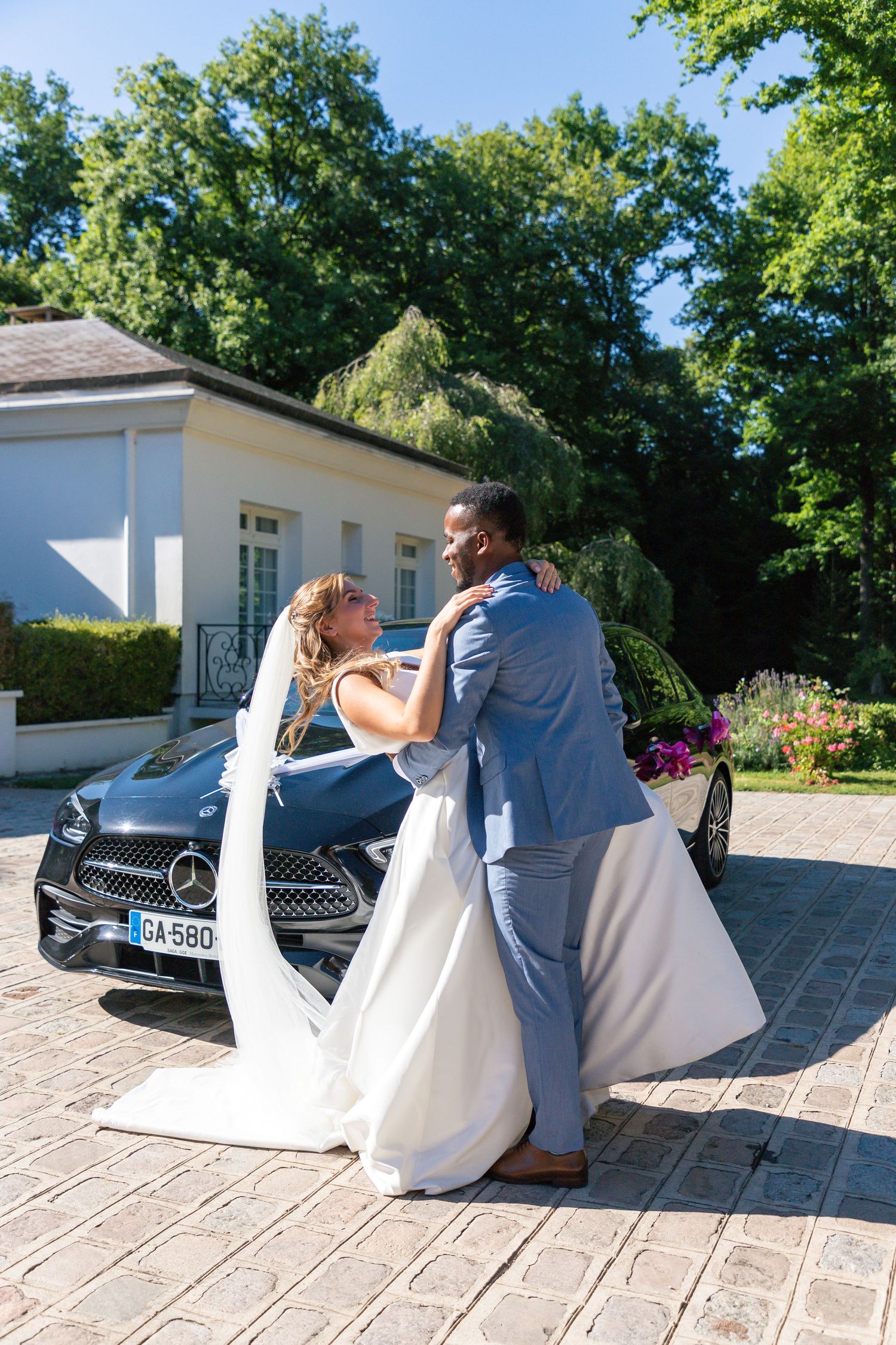 Newlyweds embracing by luxury Mercedes car outside white building.