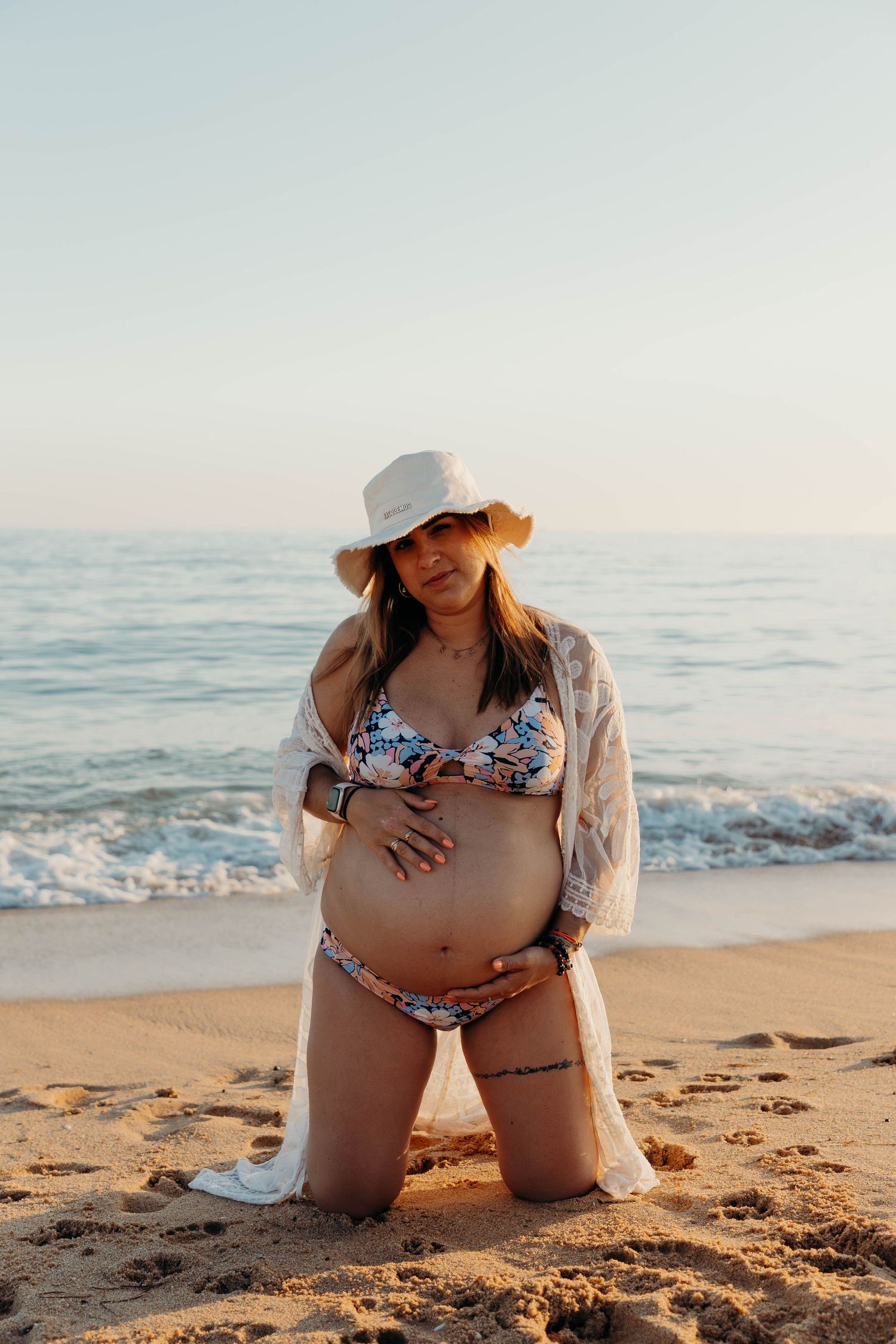 Pregnant woman in swimsuit and lace coverup poses on sunny beach.