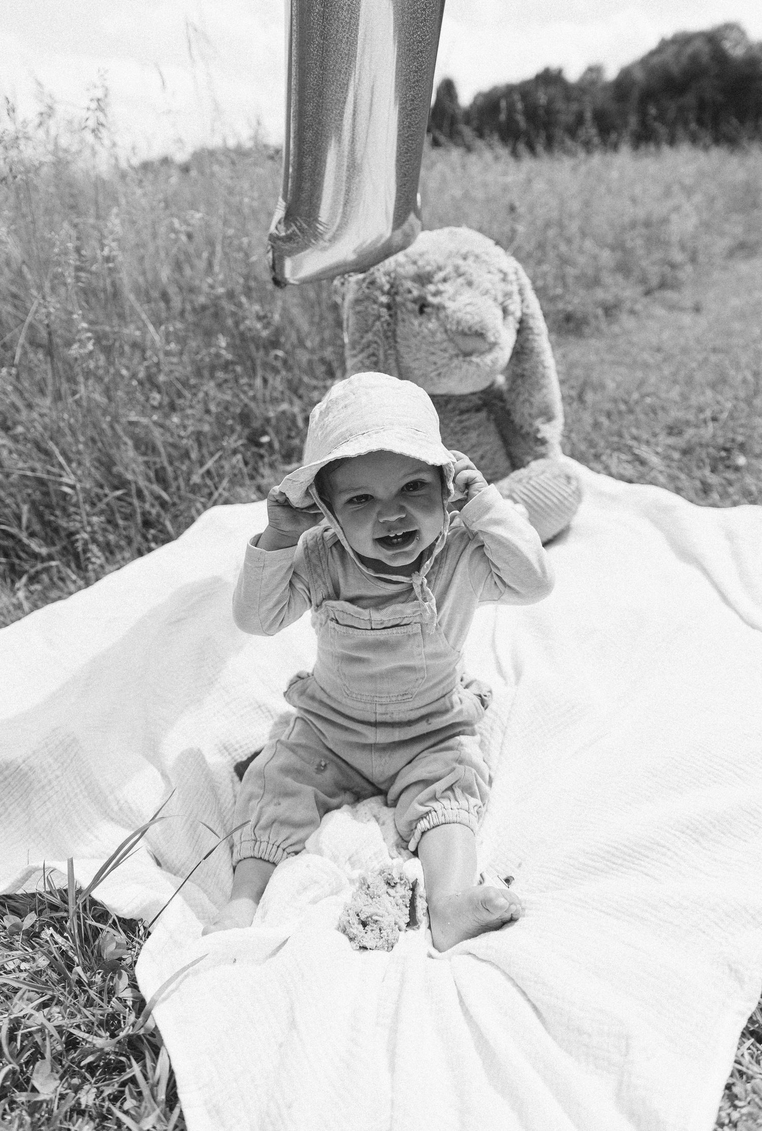 Black and white photo of baby in overalls playing on blanket outdoors.