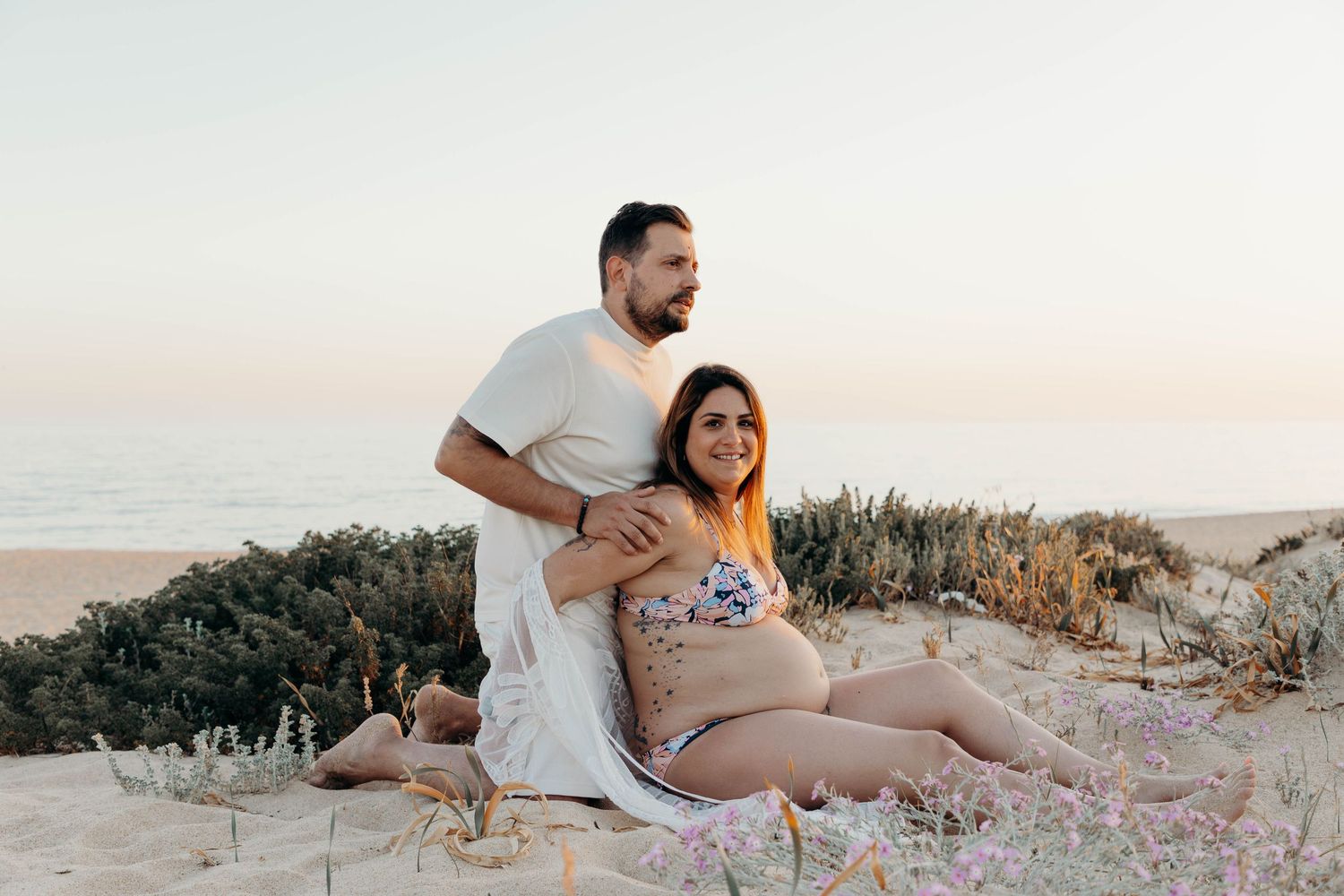 Expectant couple sitting together on sand dunes during a romantic maternity photoshoot.